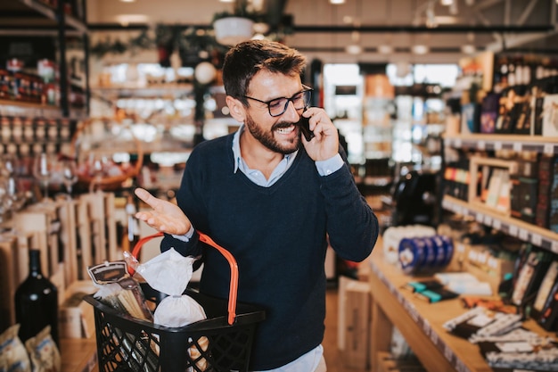 Heureux homme d'âge moyen achetant des aliments et des boissons sains dans un supermarché moderne et parlant au téléphone.