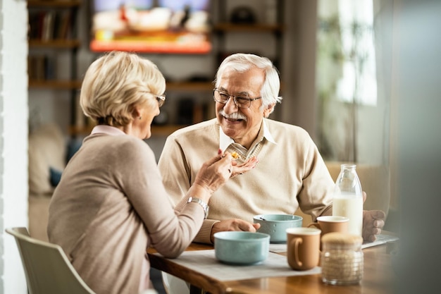 Heureux homme âgé s'amusant pendant que sa femme le nourrit pendant la table à manger du petit-déjeuner