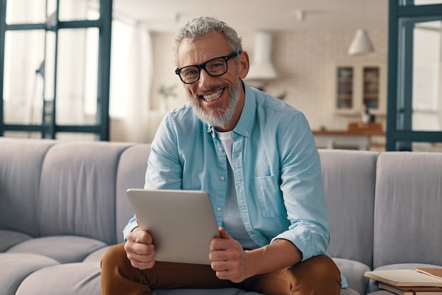 Heureux homme âgé utilisant une tablette numérique et regardant la caméra assis sur le canapé à la maison