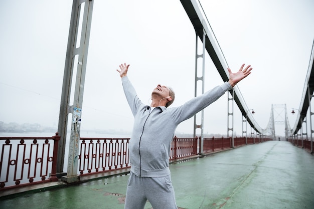 Heureux homme âgé en vêtements de sport gris debout sur le pont avec les mains en levant