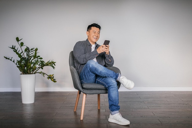 Heureux homme asiatique adulte tapant sur un smartphone assis dans un fauteuil dans un espace libre intérieur de salon minimaliste