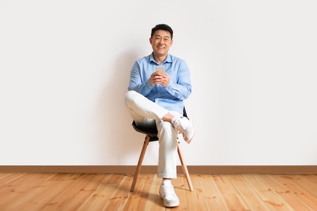 Heureux homme asiatique d'âge moyen en chemise utilisant un téléphone portable et des réseaux dans les médias sociaux assis sur