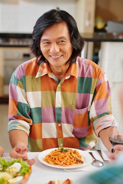 Heureux homme asiatique d'âge moyen mangeant une assiette de délicieux spaghettis et écoutant des amis lors d'un dîner de fête