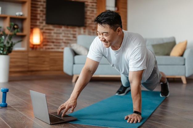 Heureux homme asiatique d'âge moyen près d'un ordinateur portable debout dans une planche faisant de l'exercice à l'intérieur du salon à la maison