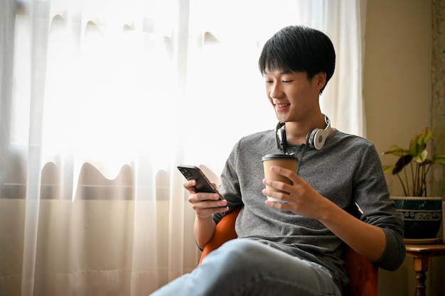 Heureux homme asiatique dans des vêtements à la mode en utilisant son téléphone tout en sirotant un café dans le café