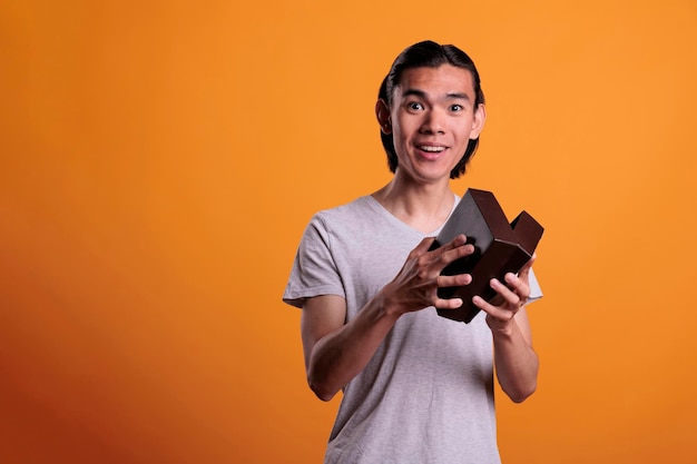 Heureux homme asiatique ouvrant une boîte-cadeau, déballant une surprise d'anniversaire. Souriant jeune personne déballant le cadeau, tenant un petit colis de livraison en carton avec une expression faciale excitée