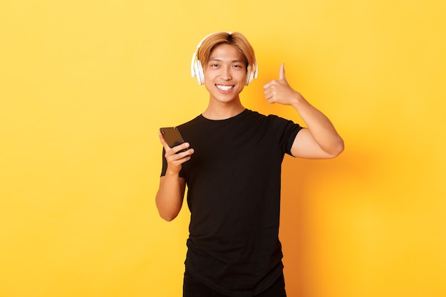 Heureux homme asiatique satisfait aime la musique ou le podcast, montrant le pouce vers le haut en approbation, tenant un téléphone portable, mur jaune debout