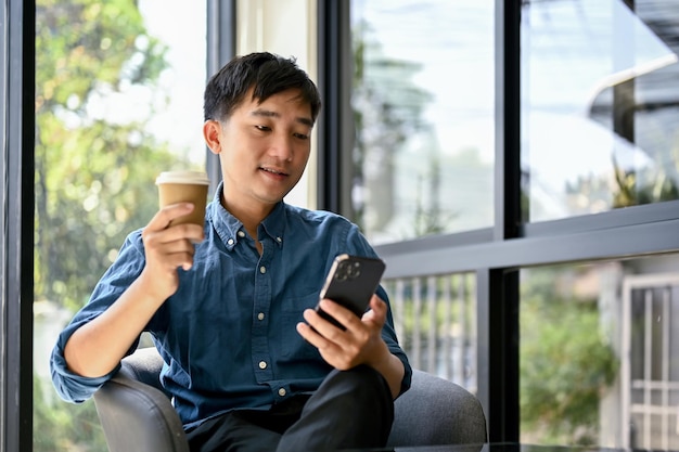 Heureux homme asiatique utilisant son smartphone et dégustant son café dans un café