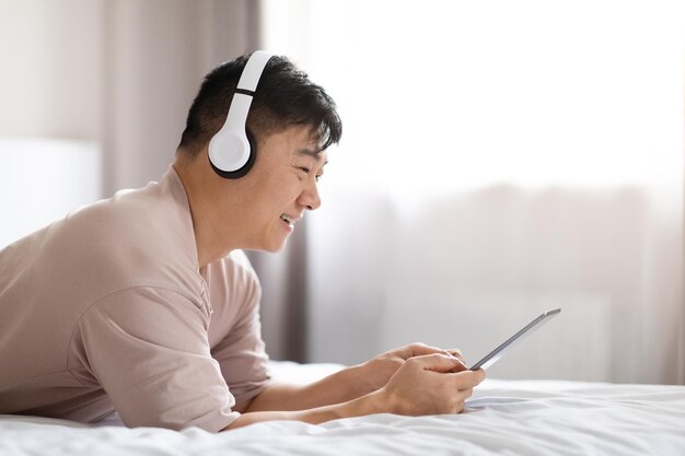 Heureux homme asiatique utilisant une tablette numérique et un casque au lit