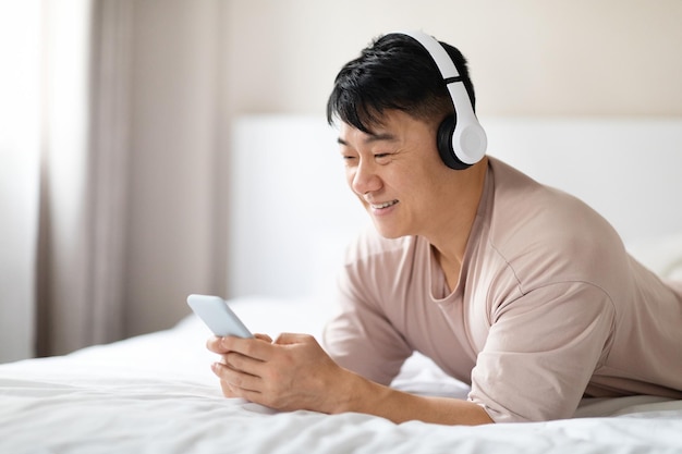 Heureux homme asiatique utilisant un téléphone portable et un casque au lit