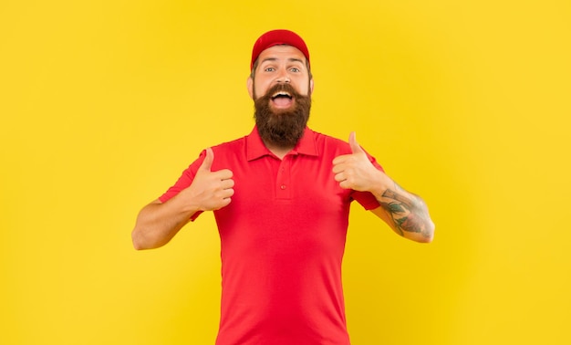 Heureux homme barbu en casquette rouge décontractée et t-shirt donnant un double pouce sur fond jaune, livreur