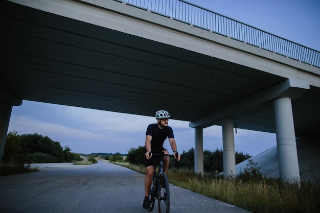 Heureux homme barbu dans un casque de protection, des lunettes en miroir et des vêtements de sport faisant du vélo sur une route goudronnée à l'extérieur de la ville le soir