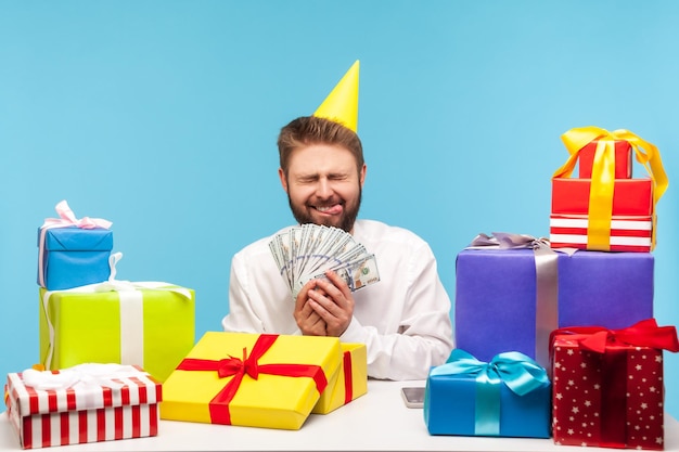 Heureux homme barbu excité en chapeau de fête se réjouissant sincèrement montrant la langue et tenant de l'argent en dollars, déballant le cadeau, recevant un bonus en espèces. Tourné en studio intérieur isolé sur fond bleu