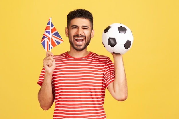 Heureux homme barbu excité en t-shirt rayé souriant tenant par la main le drapeau du Royaume-Uni et le ballon de football, soutenant l'équipe préférée du championnat. Tourné en studio intérieur isolé sur fond jaune