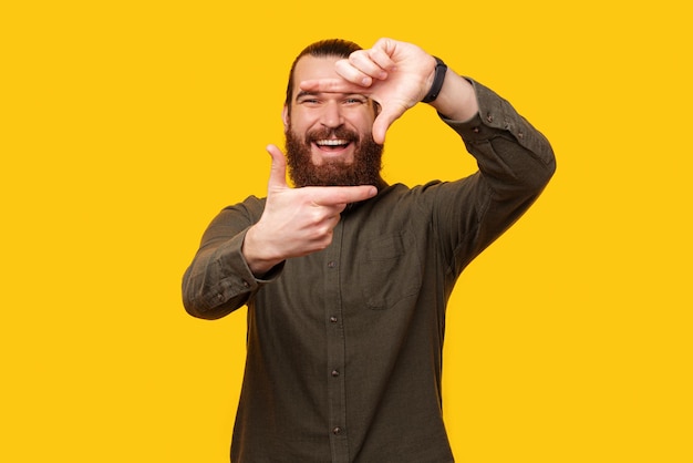 Heureux homme barbu fait un cadre d'appareil photo avec ses doigts tout en souriant à la caméra Studio tourné sur fond jaune