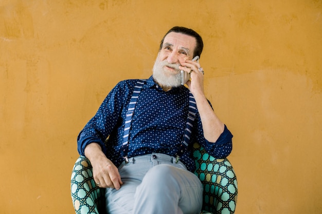 Heureux homme barbu gris à la mode moderne en chemise bleu foncé élégant, bretelles et pantalon gris, assis sur la chaise sur fond jaune et téléphone parlant