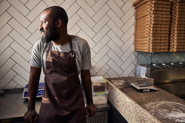 Heureux homme barbu en tablier se tient debout et se détend à table après avoir fait de la pâte et l'avoir cuite