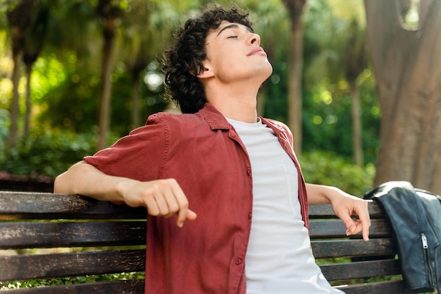 Heureux homme bouclé relaxant les yeux fermés assis sur le banc du parc