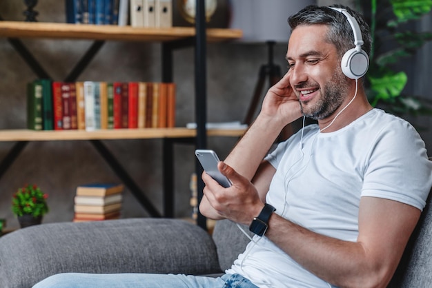 Heureux homme caucasien écoutant sa chanson préférée sur son téléphone portable avec un casque sur le canapé à la maison