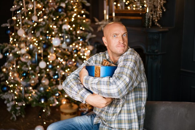 Heureux homme chauve dans une chemise tenant une boîte bleue avec un cadeau sur le fond d'un arbre de Noël