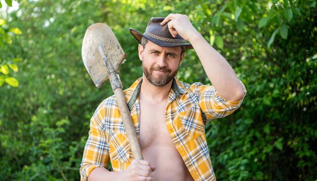 Heureux homme cowboy basculant chapeau de cowboy tenant une pelle Cowboy souriant dans le fond naturel du jardin