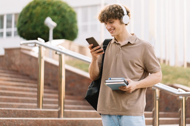 Heureux homme étudiant attrayant avec sac à dos et livres regardant le téléphone près de l'université