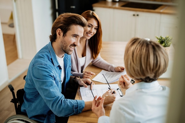 Heureux homme en fauteuil roulant et sa femme discutant avec un agent immobilier tout en analysant des plans de logement lors d'une réunion à la maison