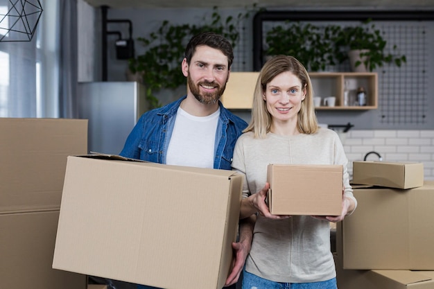 Heureux homme et femme de famille ensemble dans un nouvel appartement regardant la caméra et souriant tenant des boîtes en carton pendaison de crémaillère