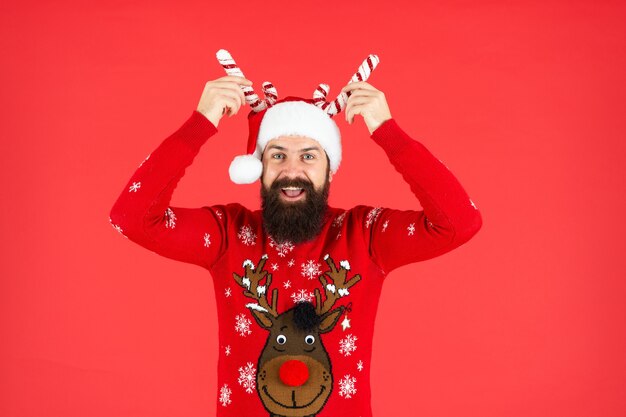 Heureux homme hipster brutal en bonnet de noel avec décoration de fête sur fond rouge, noël.