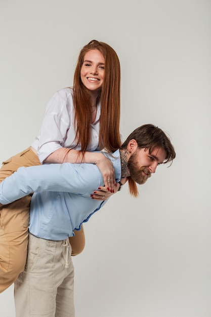 Heureux homme hipster portant sa petite amie sur son dos sur un fond gris Beau couple de mode drôle
