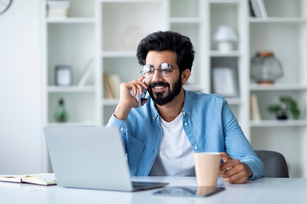 Heureux homme indien ayant une conversation téléphonique et buvant du café au bureau à domicile