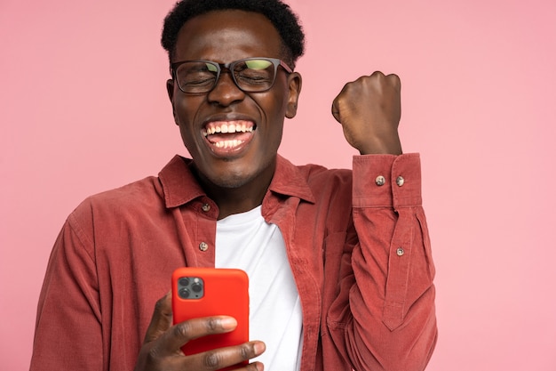 Heureux homme millénaire noir tenir téléphone mobile, faisant le geste gagnant sur rose