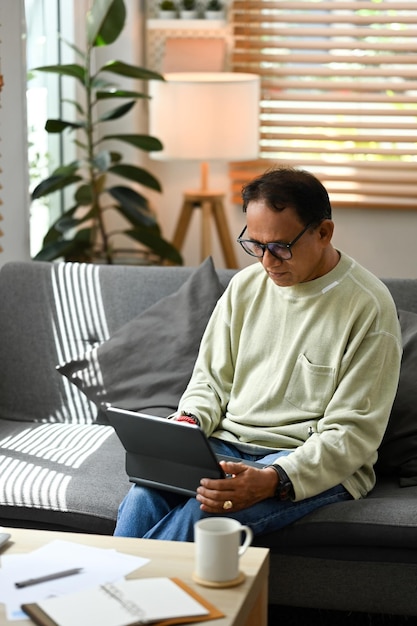 Heureux homme mûr assis sur un canapé et vérifiant ses e-mails en lisant les actualités en ligne sur une tablette Concept de technologie pour personnes âgées
