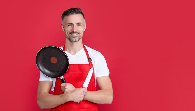 Heureux homme mûr chef en tablier avec poêle et couteau sur fond rouge