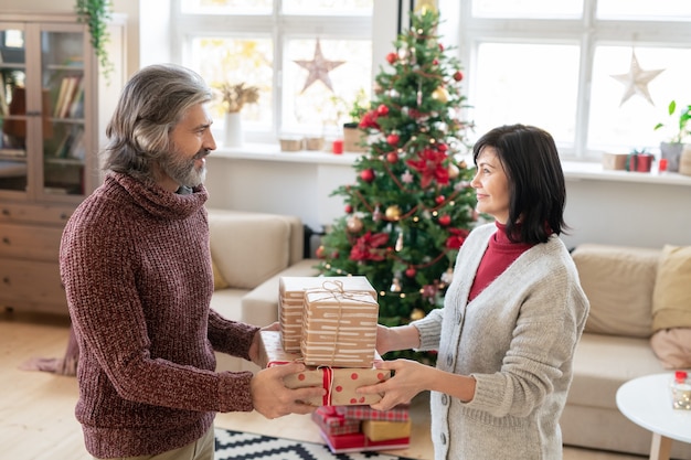 Heureux homme mûr passant trois coffrets cadeaux à sa jolie femme tout en la regardant contre un sapin de Noël décoré et une grande fenêtre dans le salon