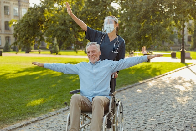 Heureux homme mûr récupérant un patient dans un fauteuil roulant lors d'une promenade avec une infirmière portant un écran facial et