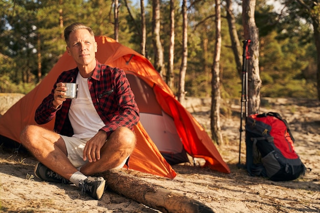 Heureux homme mûr rencontre le matin avec une tasse de boisson chaude dans la nature alors qu'il est assis près de la tente