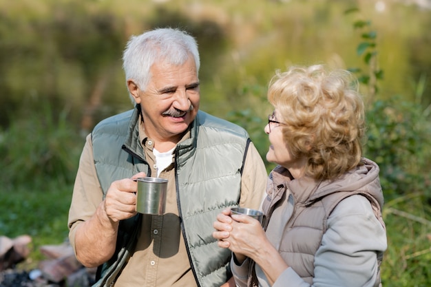 Heureux homme mûr et sa femme avec des tasses touristiques se regardant pendant une conversation dans un environnement naturel