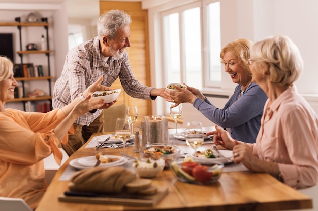 Heureux homme mûr saumurant de la nourriture à table tout en déjeunant avec ses amis à la maison