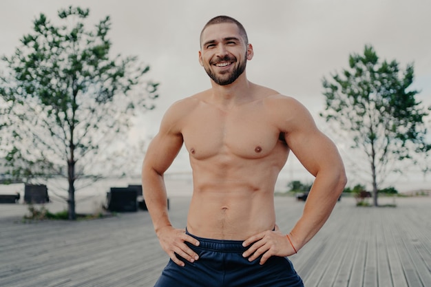 Heureux homme musclé avec poils pose en toute confiance les mains sur la taille bénéficie d'un cadre extérieur Fitness état d'esprit positif