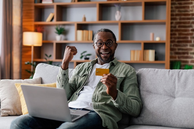 Heureux homme noir adulte excité dans des verres et décontracté se réjouit de la victoire avec ordinateur et carte de crédit