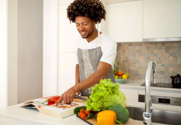 Heureux homme noir cuisiner à l'aide d'un livre de recettes en papier dans la cuisine