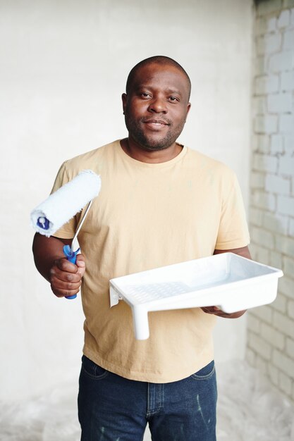 Heureux Homme D'origine Africaine Avec Rouleau à Peinture Et Récipient En Plastique Carré Blanc Avec Peinture Debout Contre Le Coin De Deux Murs