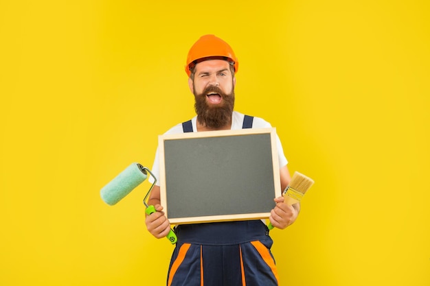 Heureux homme peintre en bâtiment en casque et vêtements de travail tenir pinceau rouleau à peinture et tableau noir avec espace de copie sur fond jaune