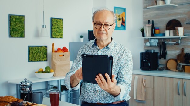 Heureux homme plus âgé dans la cuisine pendant le petit déjeuner à l'aide d'un tablet pc. Personne âgée avec tablette PC portable à l'âge de la retraite à l'aide d'applications mobiles, technologie de l'information en ligne Internet moderne avec écran tactile