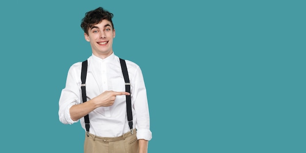 Heureux homme pointant sur fond vide et regardant la caméra avec un sourire à pleines dents. Portrait d'un jeune homme d'affaires bouclé hipster en jarretelle de chemise blanche décontractée classique. Studio tourné fond bleu isolé