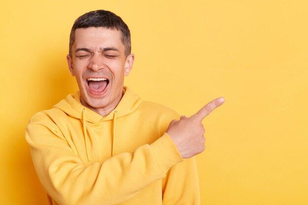 Heureux homme portant un sweat à capuche de style décontracté posant isolé sur fond jaune pointant le doigt loin avec un sourire à pleines dents sur le visage espace vide pour votre publicité en riant les yeux fermés