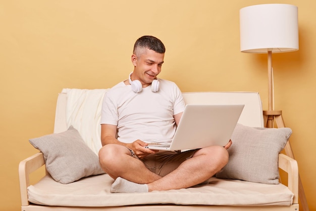 Heureux homme positif portant un t-shirt blanc et un short assis sur un canapé sur fond beige regardant un écran d'ordinateur portable regardant un film travaillant à domicile en ligne