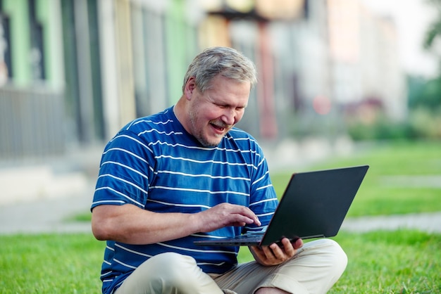 Heureux homme regardant du contenu en ligne sur un ordinateur portable et riant assis sur la pelouse de la ville