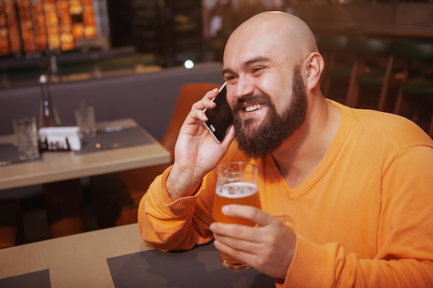 Heureux homme riant, buvant de la bière et parlant au téléphone au bar, copiez l'espace. Concept de bonne nouvelle